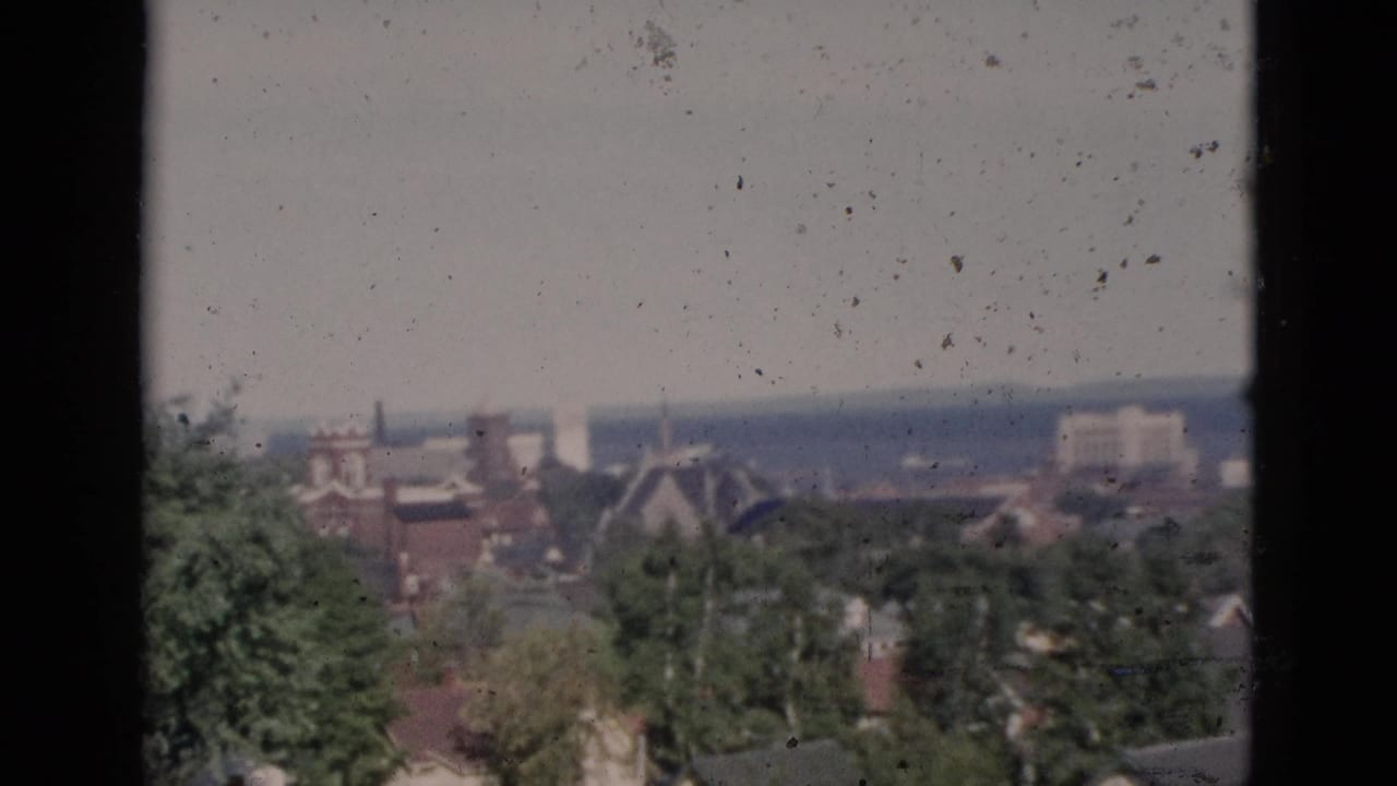 Elevated color 8mm view of a Canadian town showing a church steeple, residential rooftops, and green trees under a hazy pale sky.