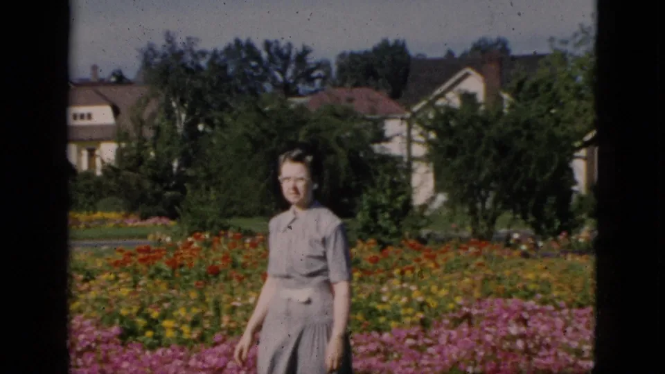 A woman wearing glasses and a gray short-sleeved top stands in a vivid summer flower garden, circa 1952 Quebec, with suburban houses and trees in the background on color 8mm film.