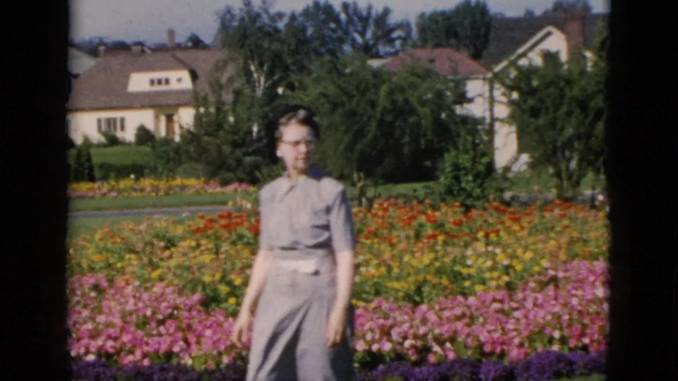 A woman in glasses among dense summer flower beds, likely Quebec circa 1952, with suburban houses and trees visible behind her on color 8mm film.