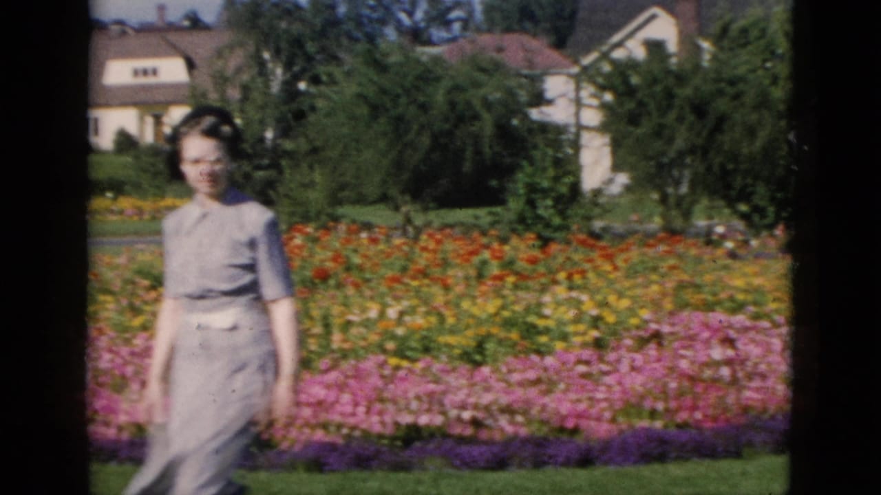 Woman in glasses and light dress walking alongside multi-colored flower beds, with residential houses and trees behind.