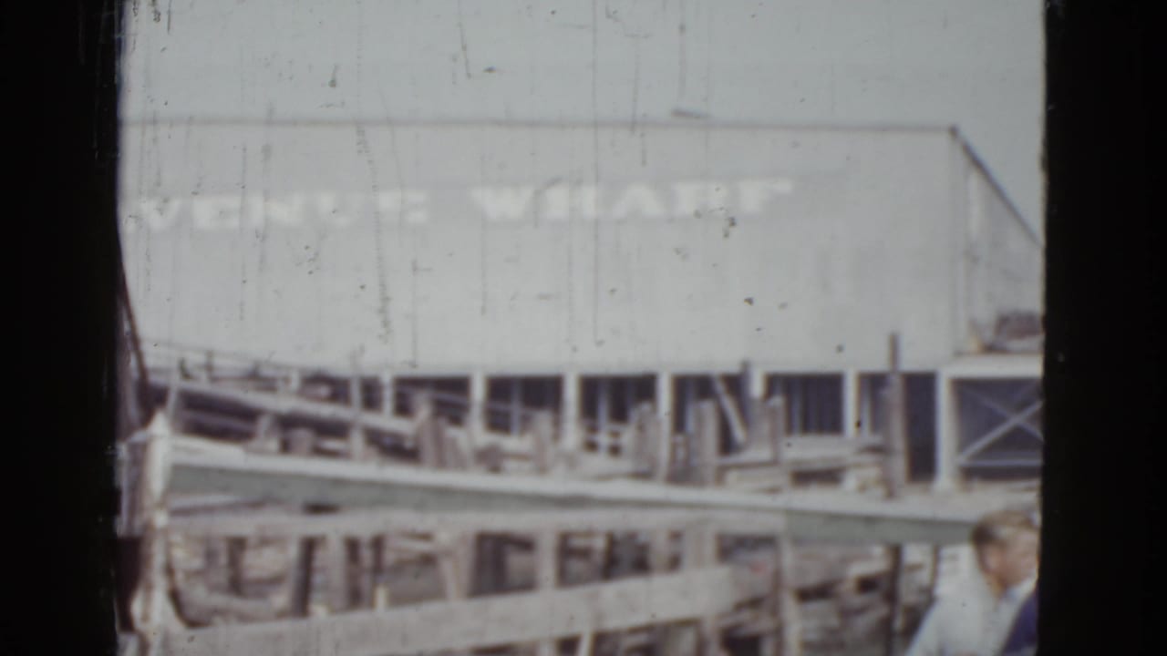 Venice Wharf building with signage, wooden dock pilings, a figure in light clothing partially visible at the right frame edge, 8mm film grain.