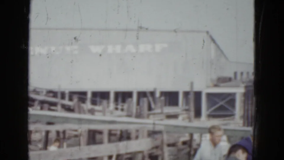 8mm home movie frame, c. 1952, New Orleans — two to three figures in period clothing at dock level beneath the Venice Wharf timber-framed building.