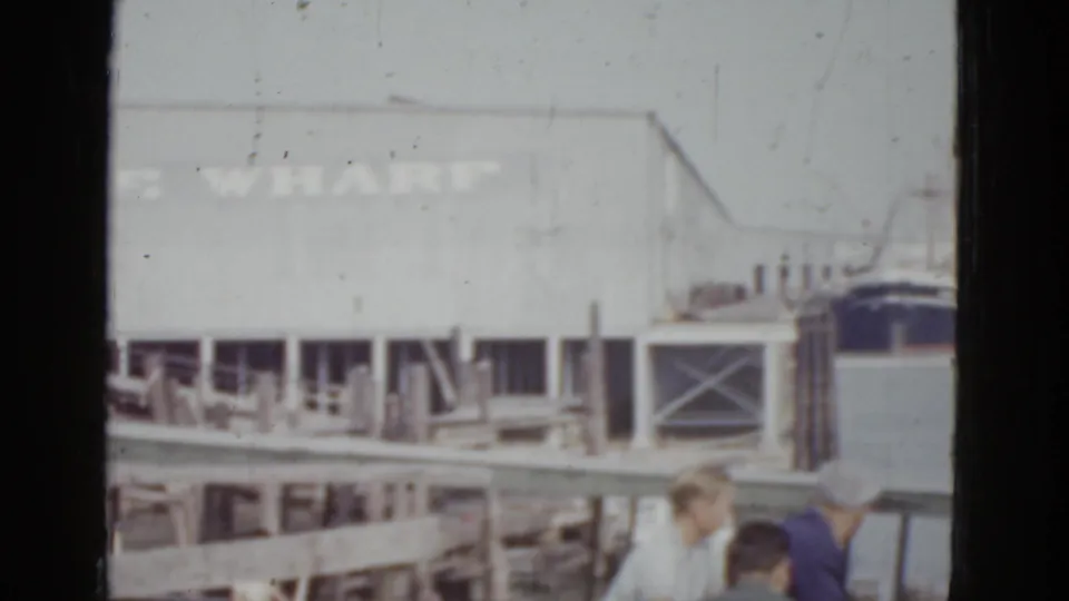 8mm home movie frame, c. 1952, New Orleans — dock-level view at Venice Wharf with two figures in period clothing, one in darker garment, partially visible at the bottom edge.