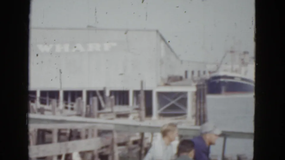 Three people in early 1950s clothing stand on a wooden dock at the New Orleans wharf, c.1952, with a large commercial vessel and a 'WHARF'-marked building behind them.