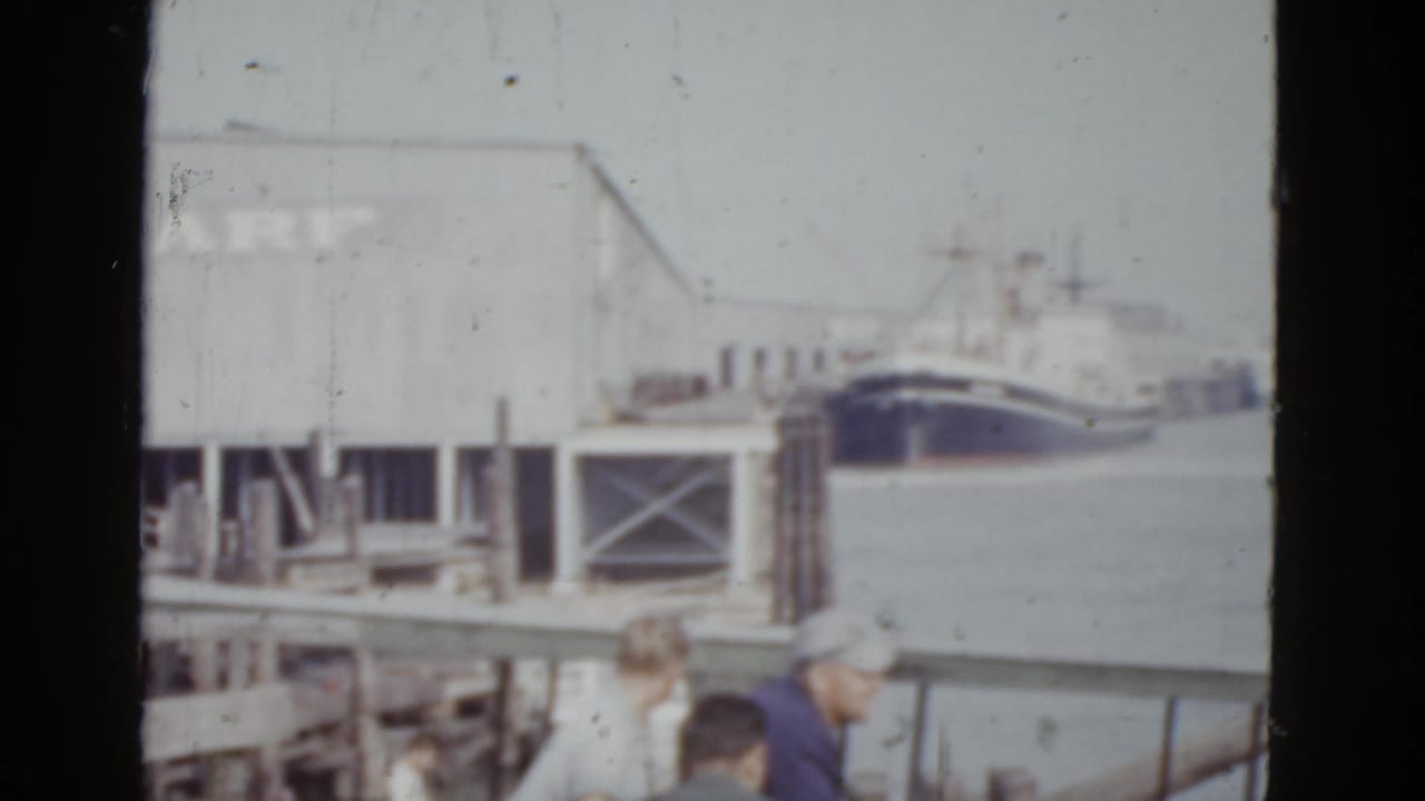 Dock scene panned slightly right; 'WHARF' sign and dark ship hull remain visible, with three figures in period clothing in the foreground.