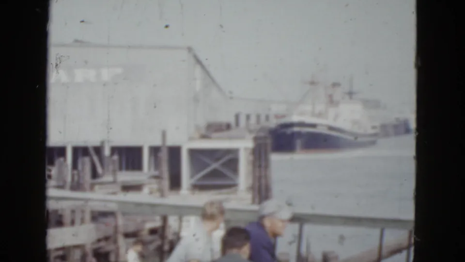 Continued pan at the New Orleans wharf, c.1952, showing the docked vessel's hull, partial 'WHARF' building signage, and a visiting group on the wooden pier.