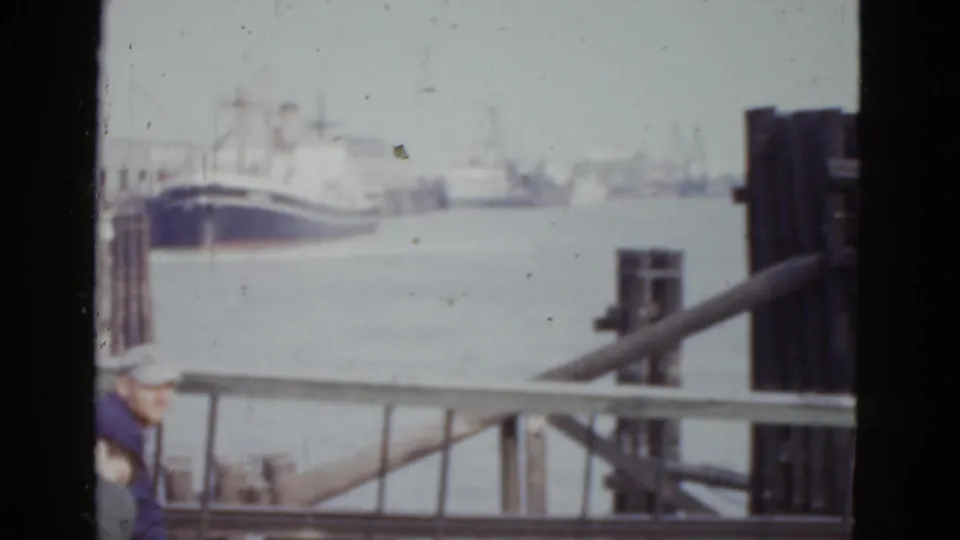 Camera pans to harbour-level view at the New Orleans waterfront, c.1952, with a moored blue-hulled boat, distant white vessels, dock railing, and a partial capped figure at lower left.
