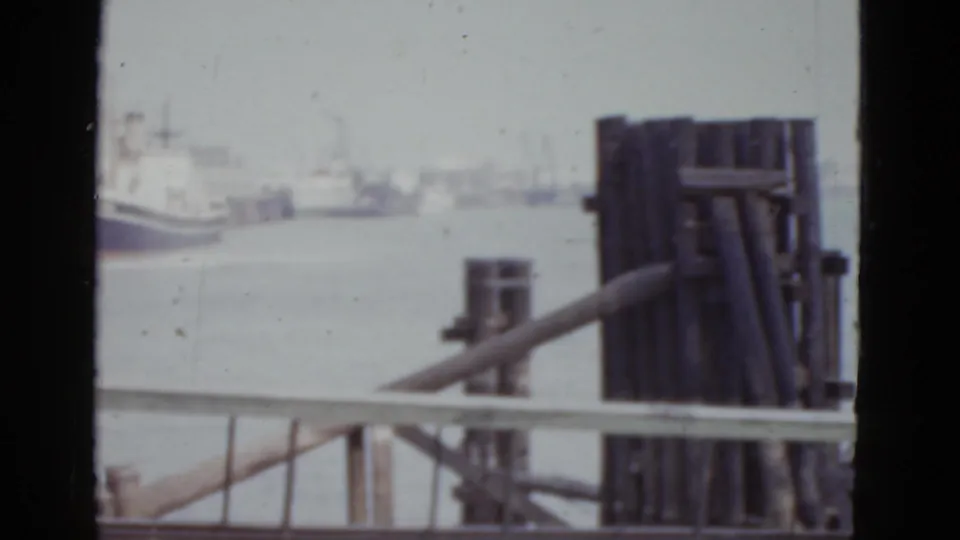 Final pan frame at the New Orleans wharf, c.1952, showing moored vessels, metal dock fencing, harbour water, and a partial capped figure at the lower edge.