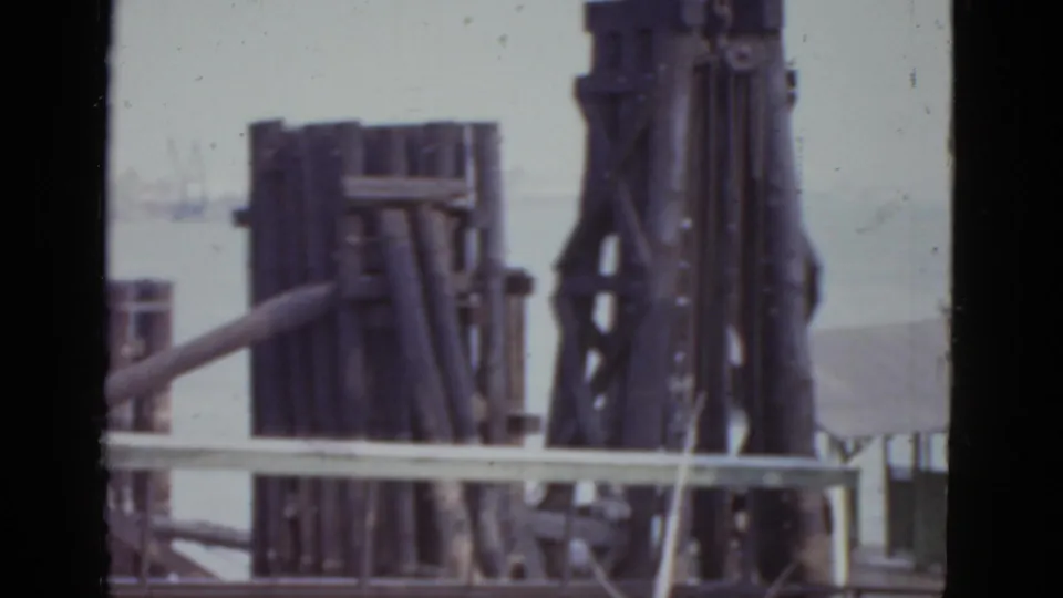Closer 1952 New Orleans harbor frame with stacked wooden pilings, vertical supports, railing, and pale water beyond.