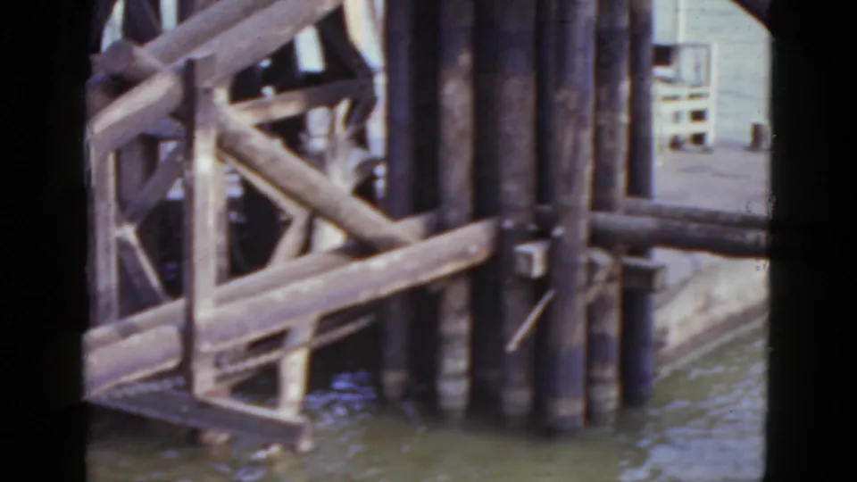 Close shot of weathered wooden dock pilings and bracing at water level with soft-focus background structure, New Orleans area, 1952 8mm.