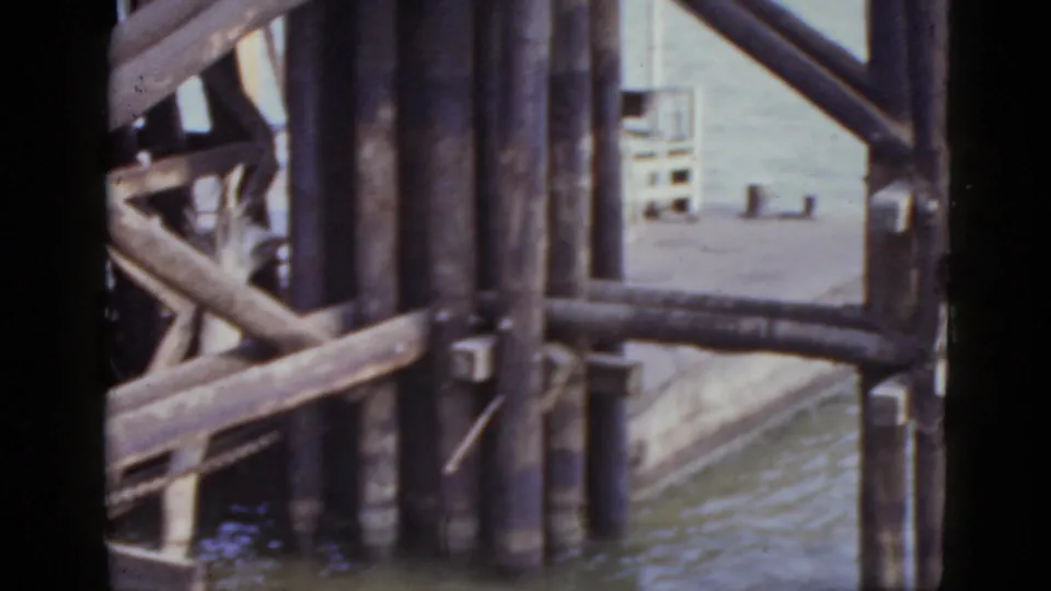 Dark wooden pilings and cross-bracing at water edge with soft background buildings, from a 1952 New Orleans waterfront 8mm home movie.