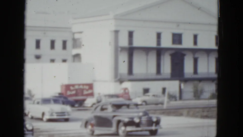 8mm color home-movie frame of early-1950s American automobiles driving on a road in front of a large white institutional building with flat pilasters, partially legible red commercial signage at left, New Orleans, 1952.