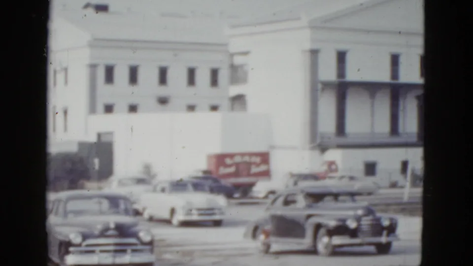 8mm color home-movie frame showing early-1950s American cars on a road in front of a large white institutional building, with a partially legible red commercial sign at right, New Orleans, 1952.