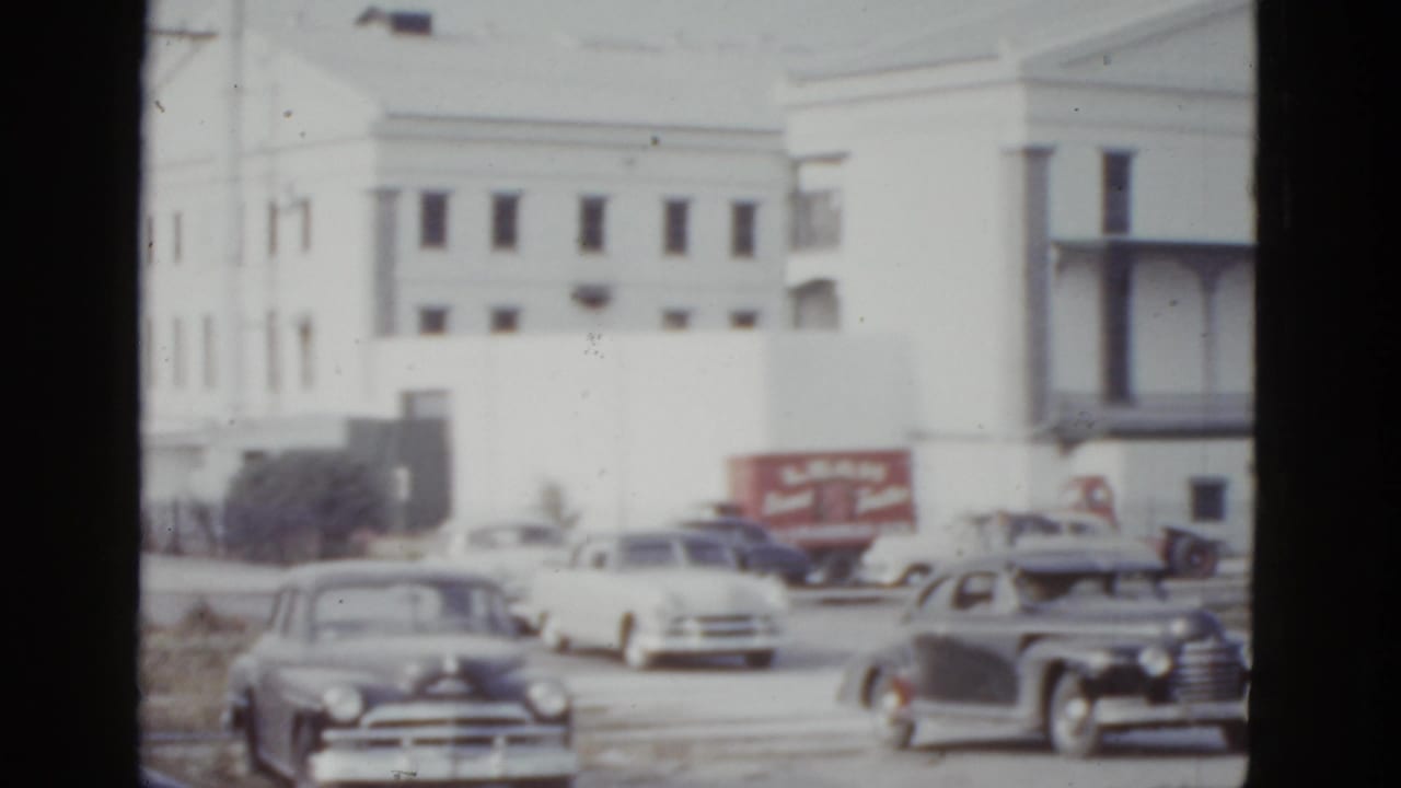 Street-level view of a large white institutional building in New Orleans, circa 1952, with rounded postwar American sedans and a red truck with commercial signage in the foreground.