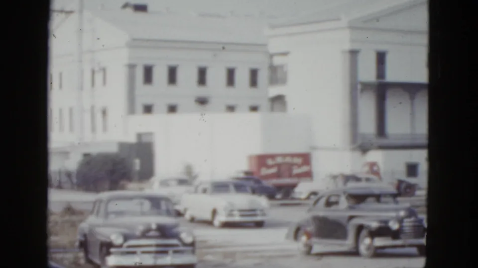 Street-level view of a large white institutional building in New Orleans, circa 1952, with rounded postwar American sedans and a red truck with commercial signage in the foreground.