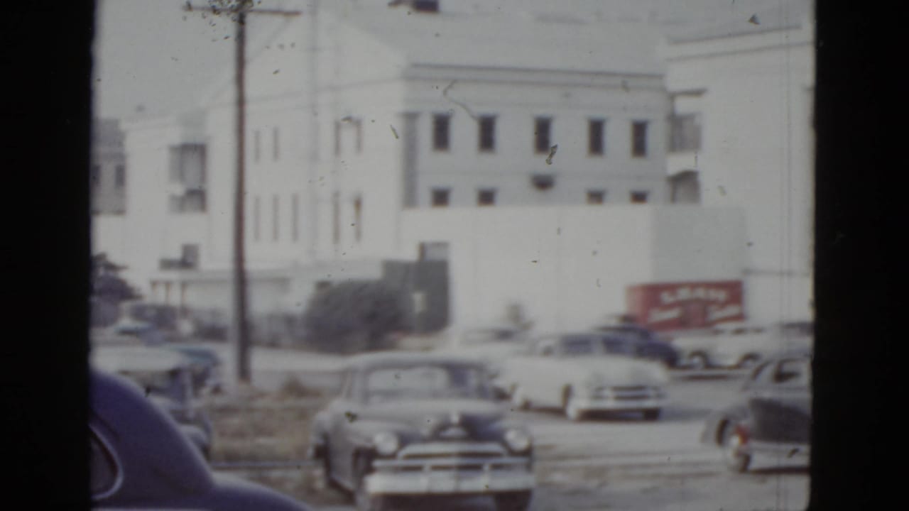 Continued view of the large white multi-story building in New Orleans, circa 1952, with early 1950s automobiles, a red truck bearing commercial signage, and a dark obstruction at the frame edge.