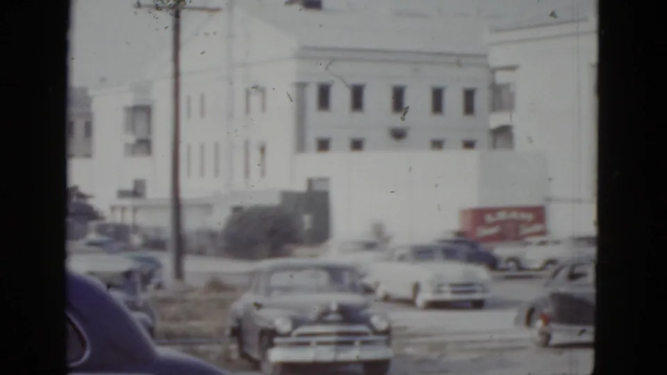 Continued view of the large white multi-story building in New Orleans, circa 1952, with early 1950s automobiles, a red truck bearing commercial signage, and a dark obstruction at the frame edge.