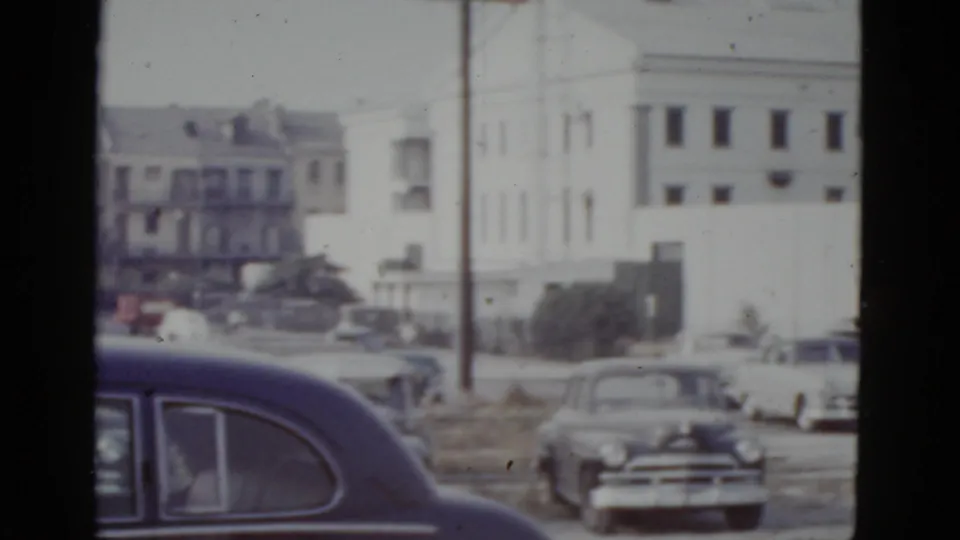Panned view in New Orleans, circa 1952, with a large dark postwar American sedan dominating the foreground and mid-century mixed-use buildings visible across an open lot.