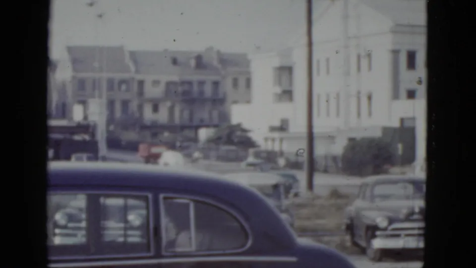 Final frame of 1952 New Orleans street showing a large dark postwar American sedan close to camera and a cluster of mid-century buildings and vehicles across an open area.