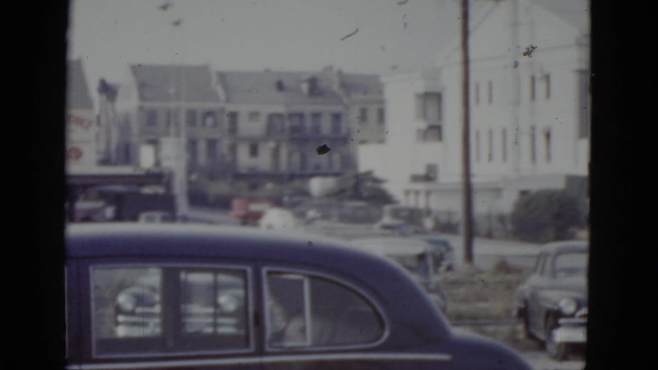 Dark blue 1940s–50s American sedan in foreground; multi-story white apartment buildings and a utility pole visible behind.