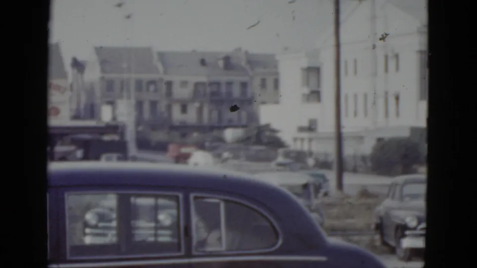 8mm home movie frame from approximately 1952 New Orleans: dark navy sedan in foreground, white multi-story residential buildings and street activity in background.