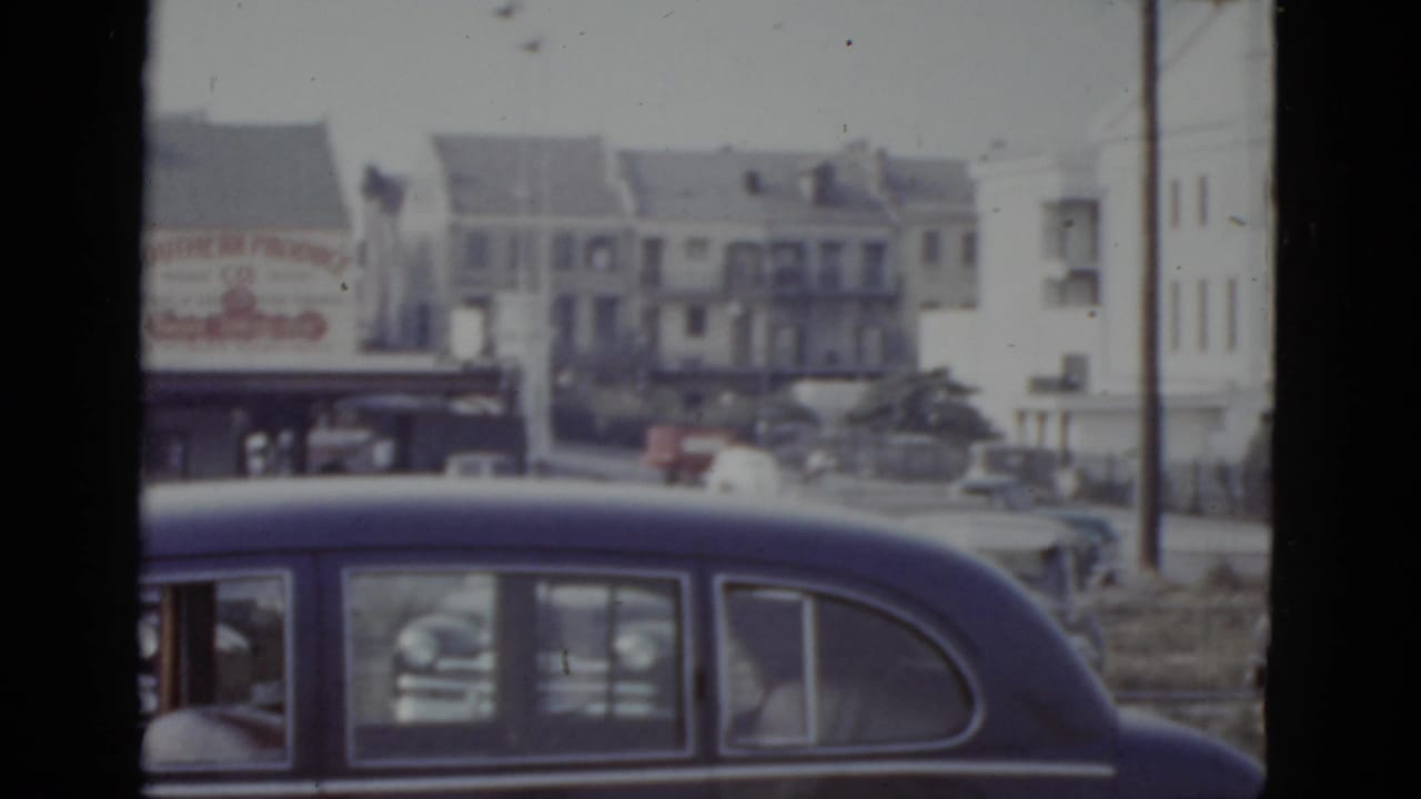 Dark blue sedan rear window in foreground; white multi-story buildings and a partially visible billboard with red lettering in background.