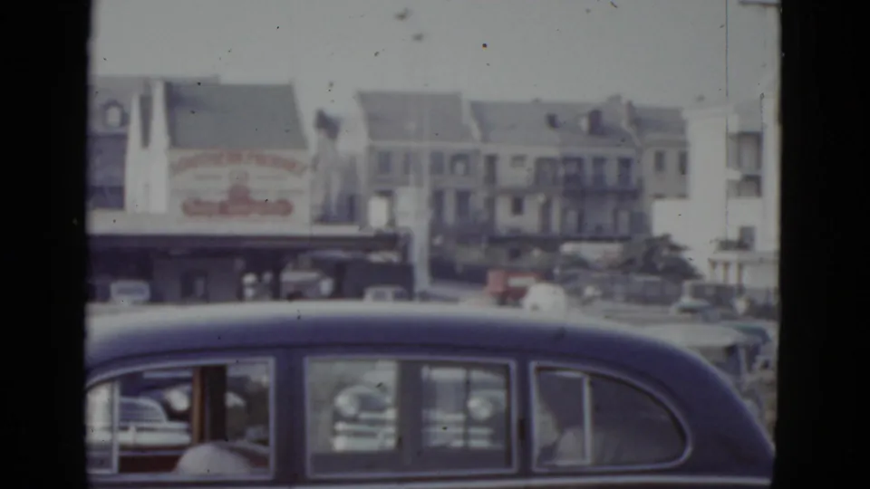 8mm home movie circa 1952, New Orleans: commercial billboard with red lettering centered in frame, white residential apartment buildings visible behind, sedan in foreground.