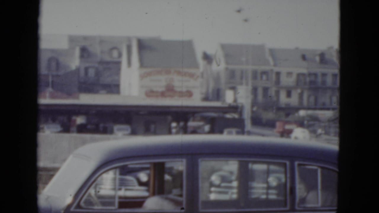 Dark American sedan in slow traffic at street level, with commercial buildings visible beyond a concrete barrier.