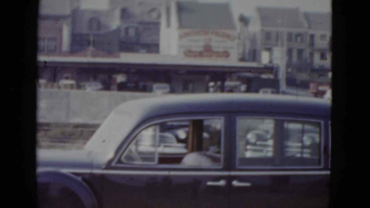 Dark sedan continues in slow traffic; a white steeple or tower is visible among background buildings.
