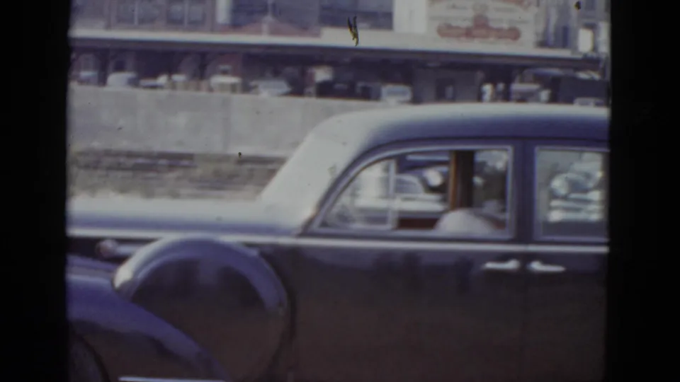 Dark American sedan beside a concrete barrier with objects on top; colorful commercial storefront signage on multi-story buildings behind; New Orleans, circa 1952 per metadata.