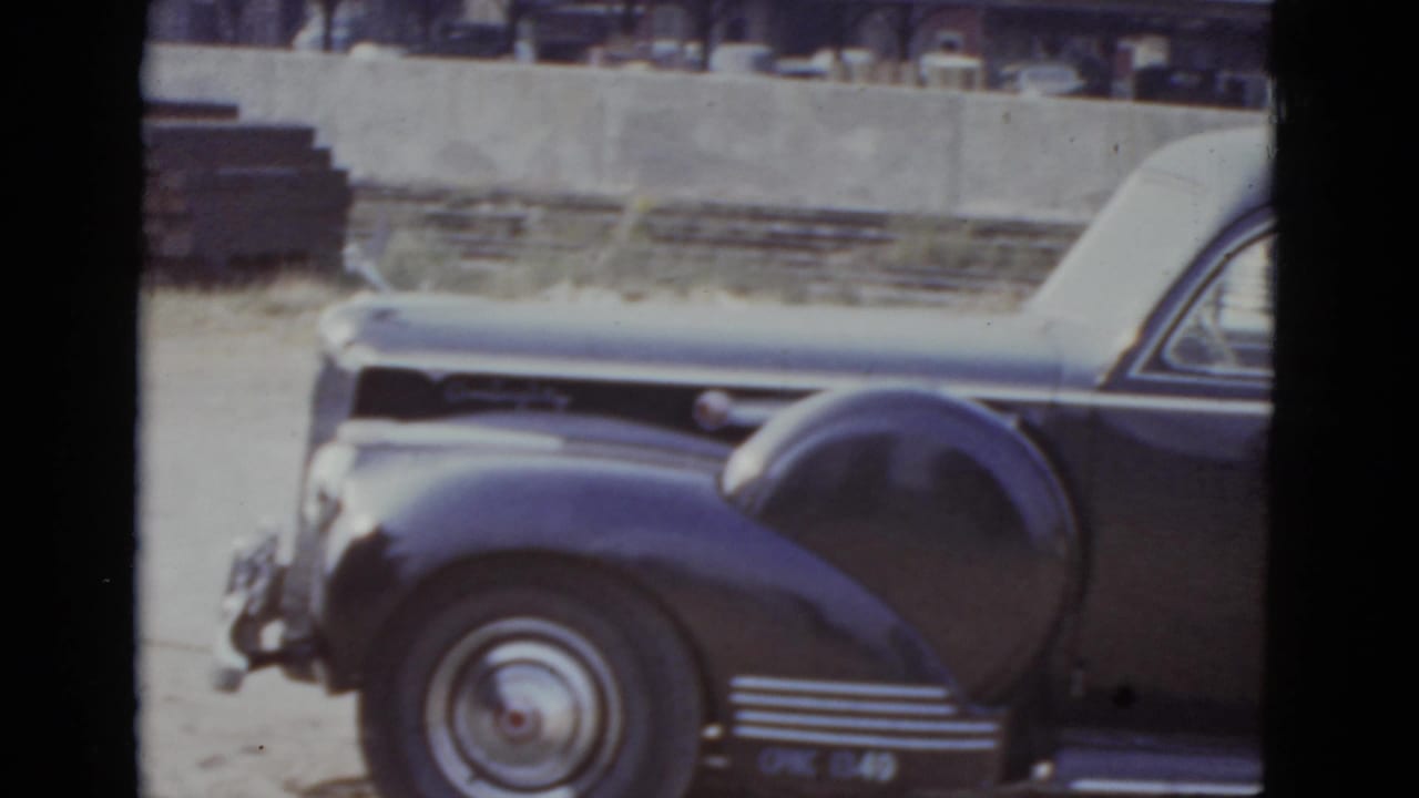 Dark-colored rounded-fender American sedan parked on a street, viewed from front-quarter angle, with a concrete wall or embankment in the background.
