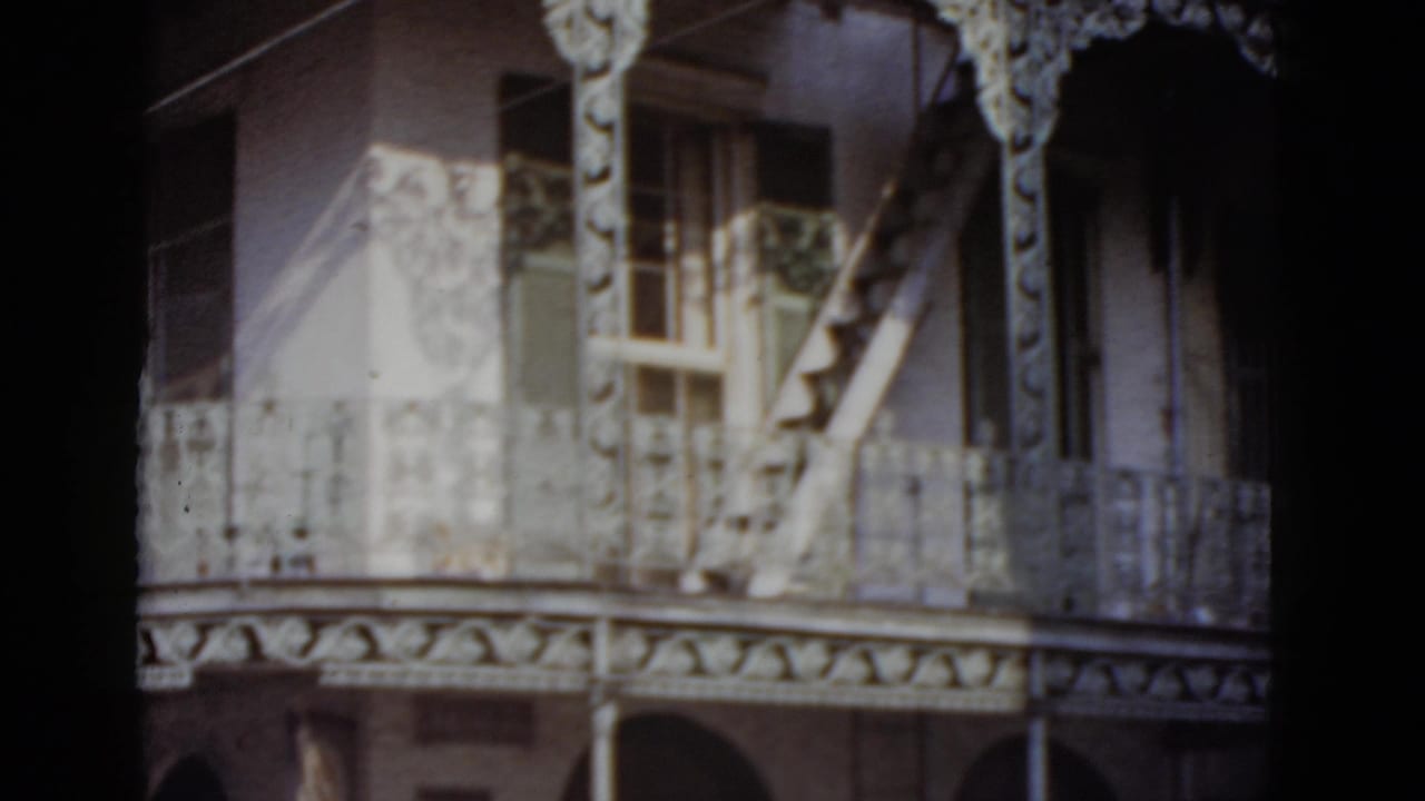 Upper floors of an ornate building with cast-iron balcony railings, decorative frieze, arched openings, and external staircase filmed on 8mm.