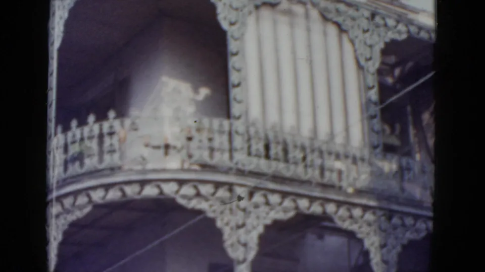 Final pan frame of New Orleans ornate ironwork residence, circa 1952, with bright light through arched openings, prominent decorative frieze, and faint vertical scratch lines on film.