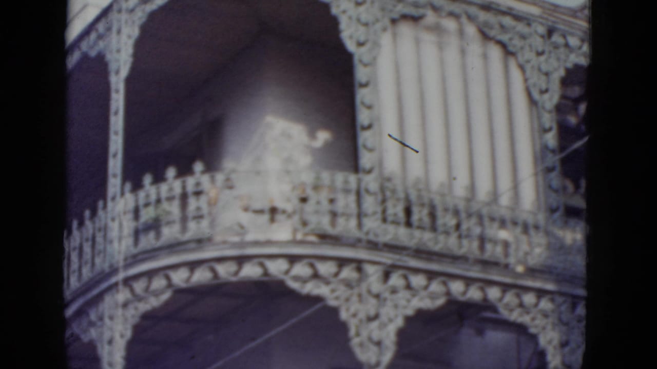 Close-up of ornate white cast-iron balcony railing and lattice panel on a multi-story New Orleans building, 8mm film.