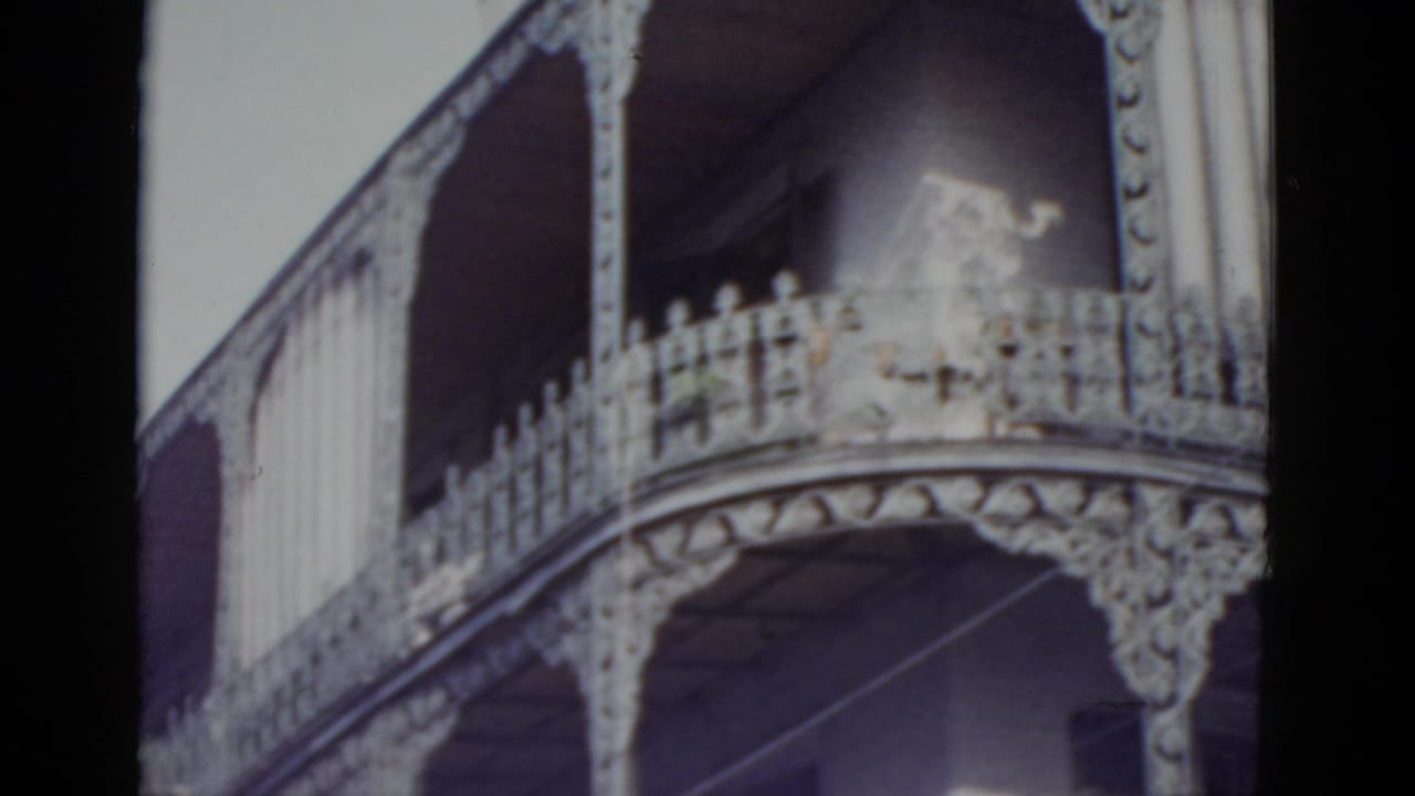 Two-level ornate iron gallery facade on a New Orleans building, camera panned slightly right, pale sky above.