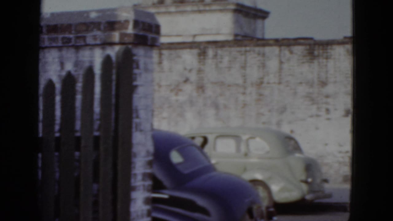 Two early-1950s cars parked behind a wooden picket fence against a weathered brick wall