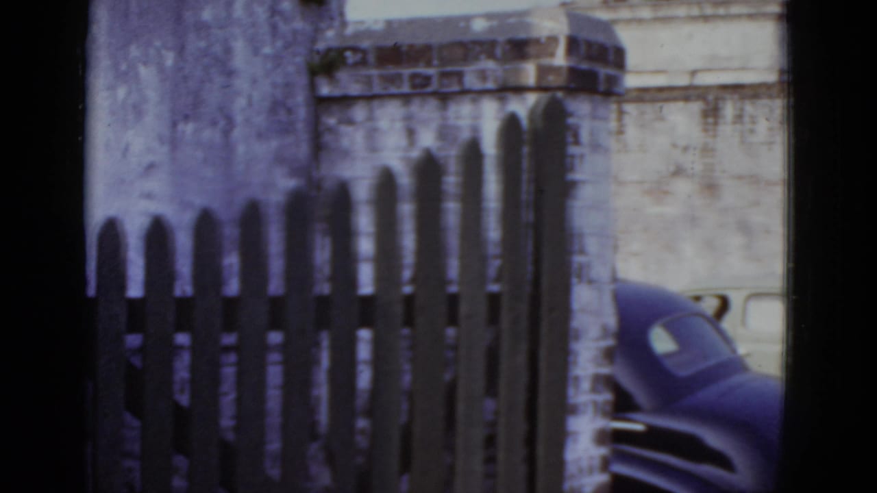 Wooden picket fence and brick pillar centered in frame; dark sedan partially visible at right edge against a brick wall