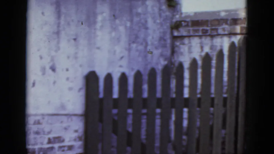 Mid-pan frame shows a close view of a wooden picket fence with a break between sections and a weathered wall behind, consistent with a New Orleans streetscape, 1952
