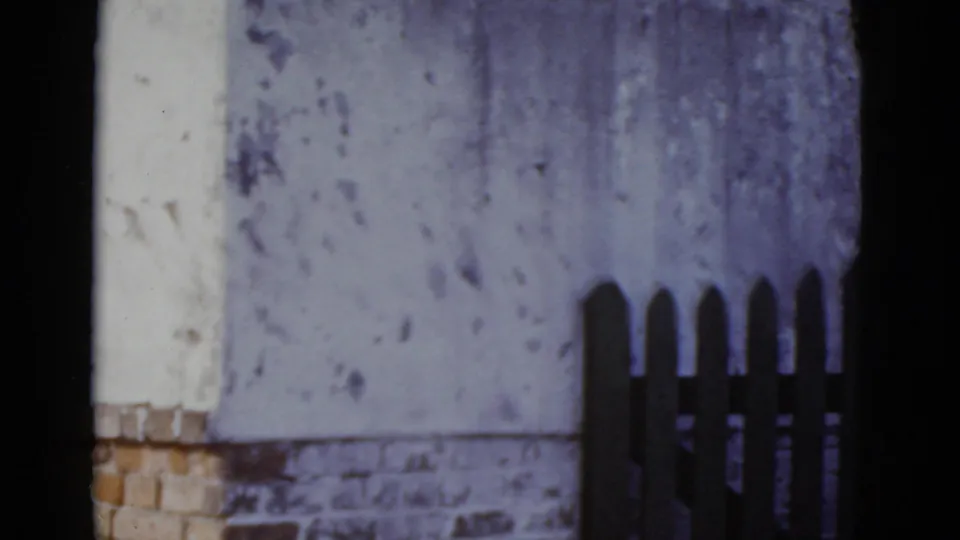 Pan ends on a weathered stucco wall with a brick lower course and the left edge of the picket fence, consistent with a New Orleans street-level exterior, 1952