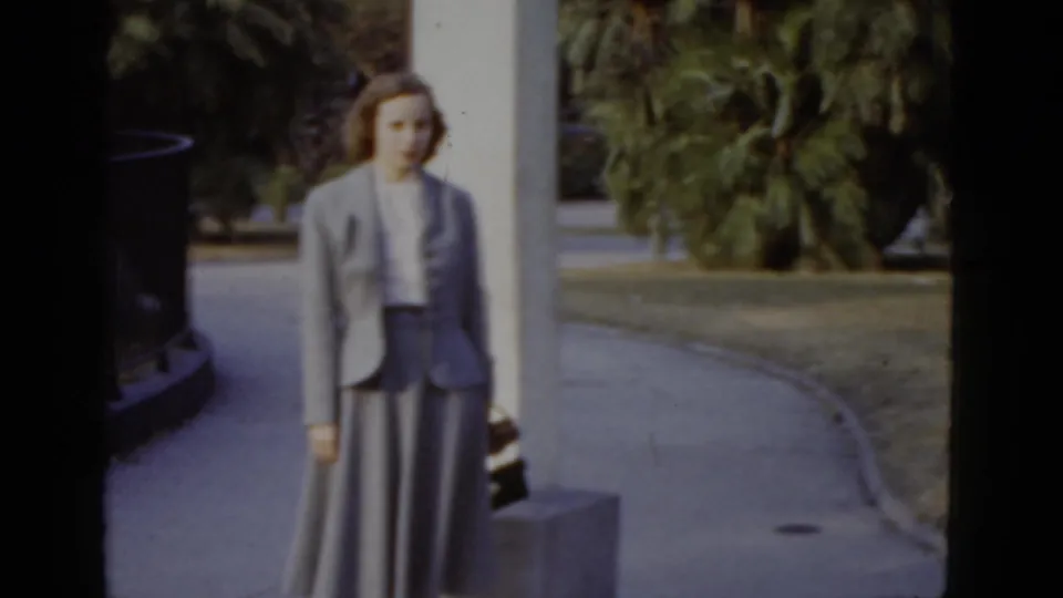 Color 8mm frame c.1952, New Orleans: woman in grey tailored suit and white blouse poses near a round concrete planter, with palm trees and a curved black iron fence in the background.