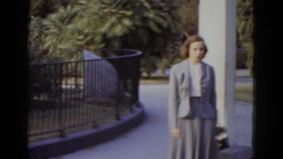 Color 8mm frame c.1952, New Orleans: woman in grey tailored suit poses beside a white column, with a curved black iron fence and lush palm trees behind her.