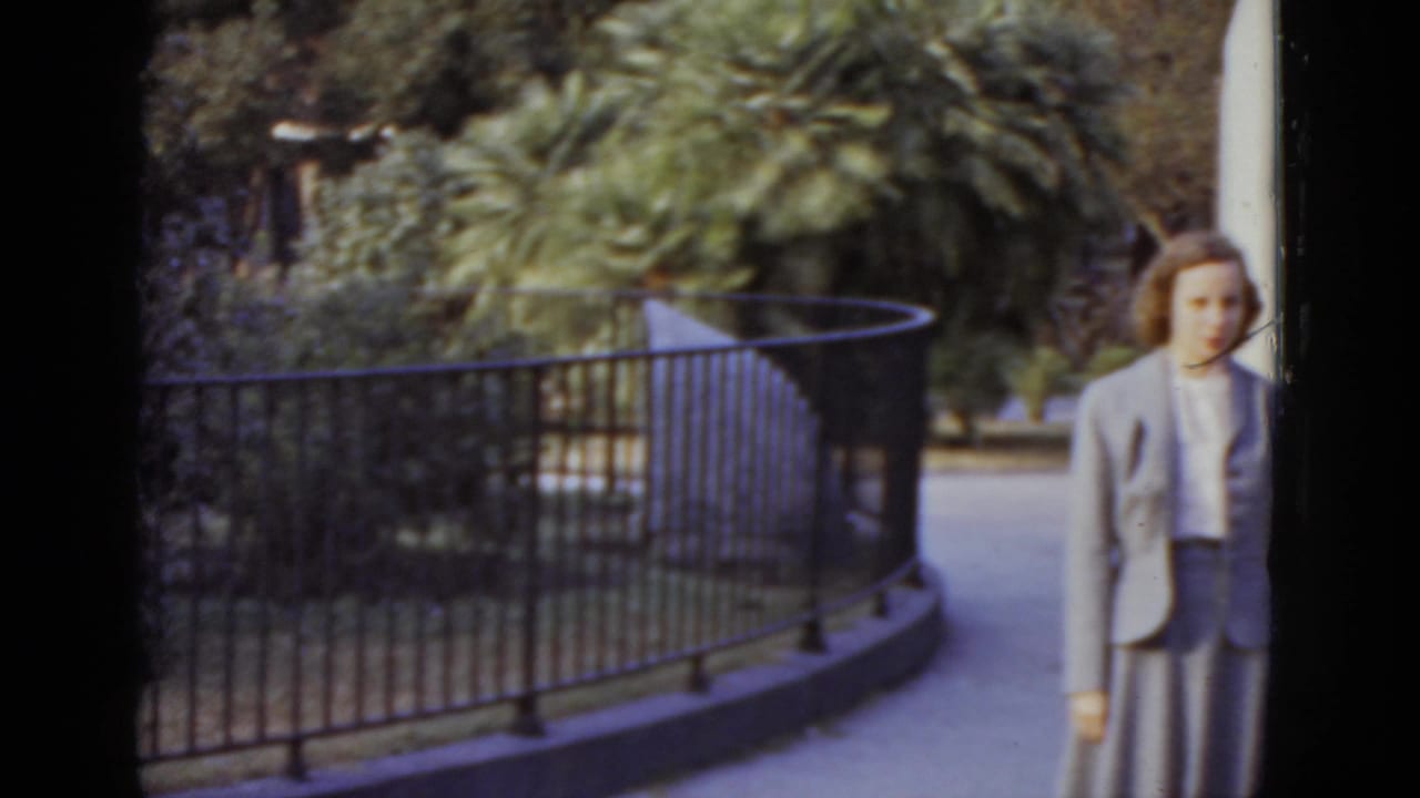 Woman in 1950s tailored suit stands near a curved iron railing in a subtropical garden, likely New Orleans, 1952; 8mm film with purple color cast.