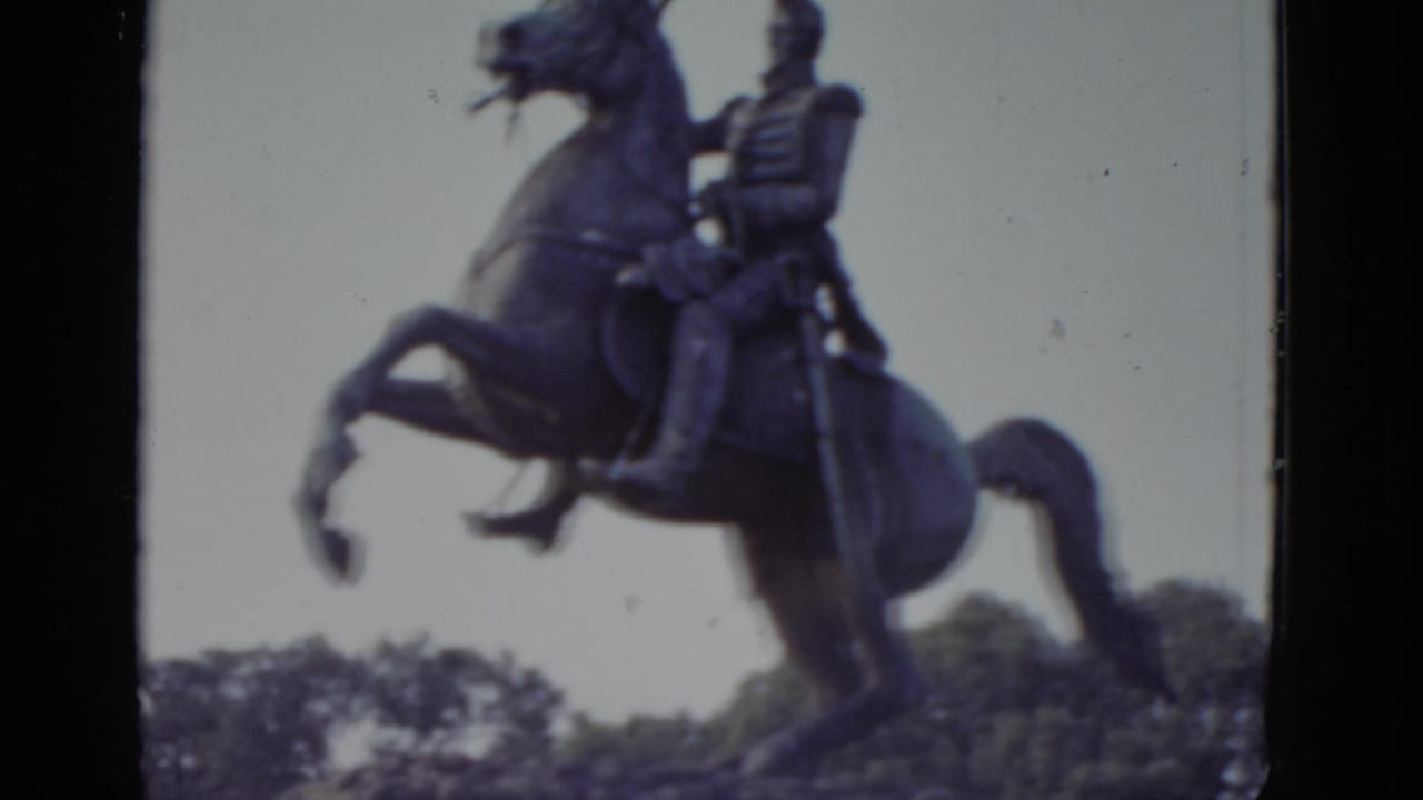 Full bronze equestrian statue on a stone pedestal showing a uniformed rider on a rearing horse, 8mm film with violet color shift.
