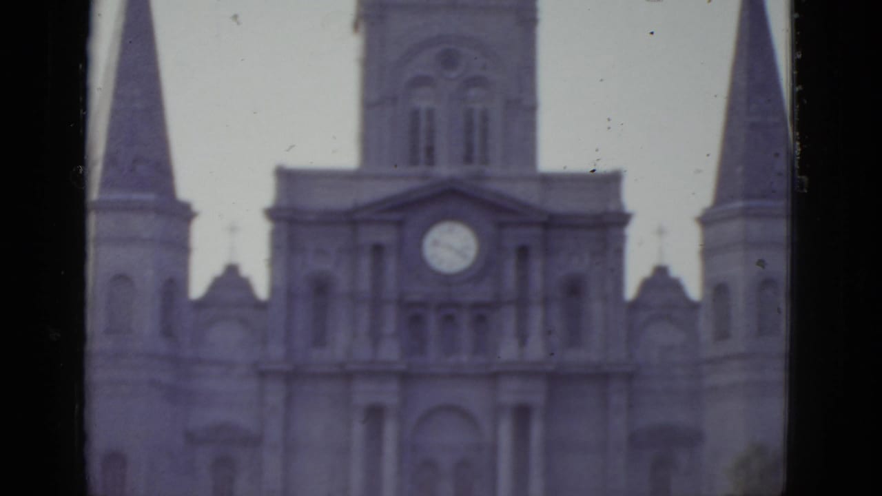 Tighter 8mm frame circa 1952 of a New Orleans Gothic cathedral's upper facade, clock face centered between decorative stonework and flanking spire bases.
