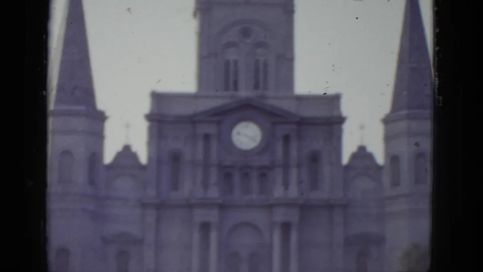 Tighter 8mm frame circa 1952 of a New Orleans Gothic cathedral's upper facade, clock face centered between decorative stonework and flanking spire bases.