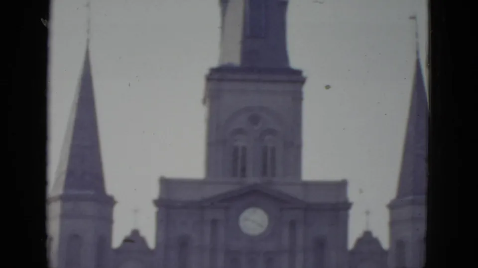 8mm tilt shot circa 1952, New Orleans Gothic cathedral — central spire and flanking towers dominate as camera angles skyward, exposure washes to pale gray.