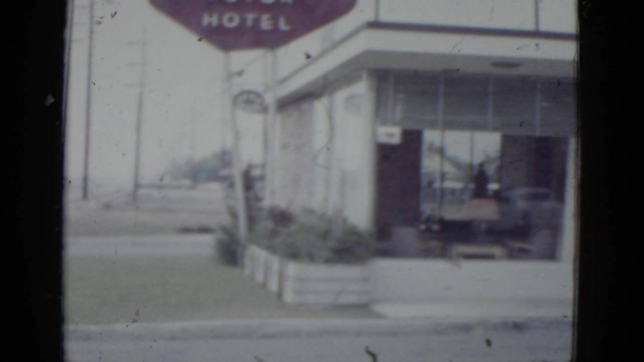 Angled close-up of a mid-century hotel exterior with a maroon HOTEL marquee sign and glass lobby windows.