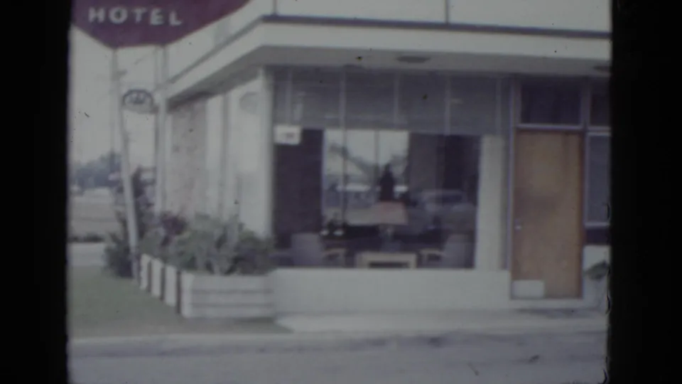 Wide shot of a flat-roofed mid-century hotel with maroon HOTEL sign, glass lobby entrance, and circular emblem on post, New Orleans area, c.1952.