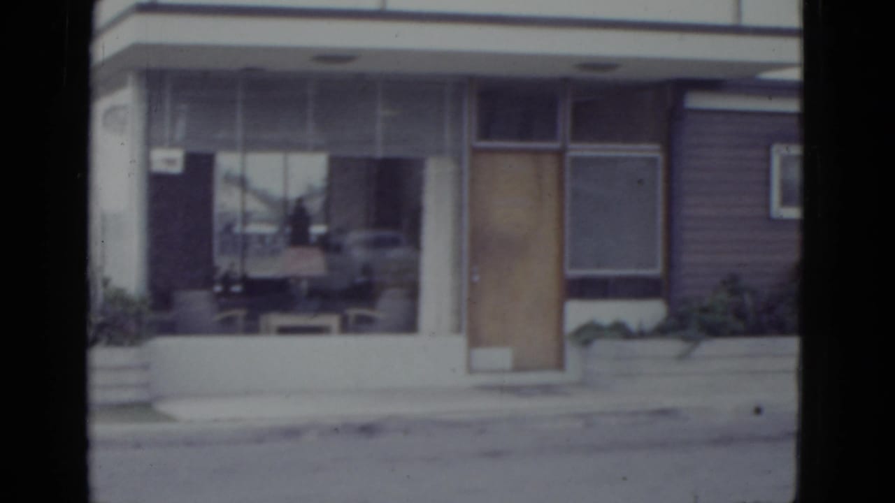 Close view of a mid-century commercial building facade with a large plate-glass window, wooden door, and horizontal reddish-brown siding.