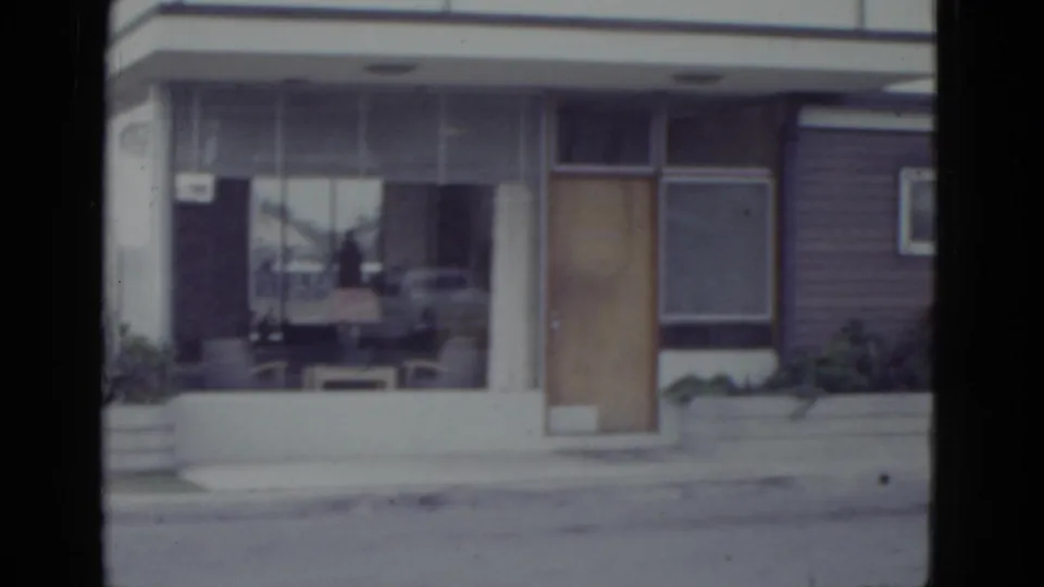 1952 New Orleans commercial building on 8mm film; large plate-glass window reflecting a period automobile, wooden entry door, and horizontal siding.